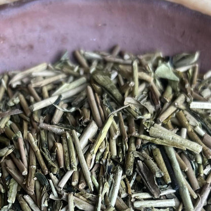 Close-up of loose dried green tea leaves piled in a dark brown ceramic bowl, with a bamboo mat background visible around the edges.