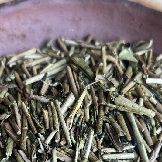 Close-up of loose dried green tea leaves piled in a dark brown ceramic bowl, with a bamboo mat background visible around the edges.