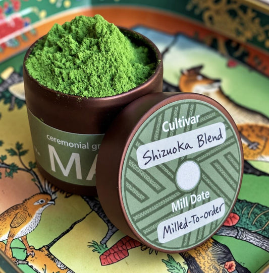 A close-up of a green ceremonial tea powder in a dark brown ceramic jar, with the lid placed in front showing printed details for 'Cultivar Shizuoka Blend'. The jar and lid rest on a decorative, colorful illustrated tray.
